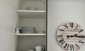 A kitchen cabinet with plates and cups of various sizes. A large clock and a neat note are hanging on the wall.