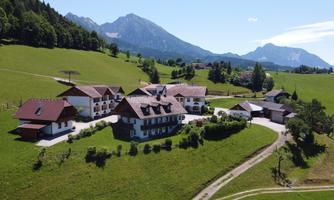 A picturesque landscape with a traditional farmhouse, surrounded by green meadows and mountains in the background. The sky is clear and blue, creating a relaxed atmosphere.