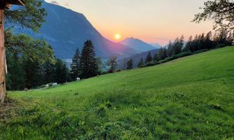 A green meadow with trees and majestic mountains in the background. The sun sets behind the mountains, coloring the sky in warm tones.