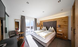 A modern hotel room with a double bed and wood paneling. There is a small desk and access to a balcony with an outside view.