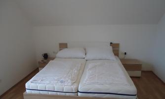 A simple bedroom with a double bed made of light wood. The walls are white and the floor is made of wood.