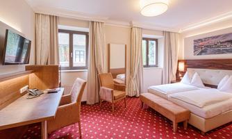 A comfortable hotel room with two beds, a desk, and a television. The windows provide daylight, and the decor is modern and inviting.