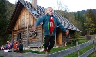 A boy happily jumps over a wooden railing in front of a rustic cabin. In the background, people can be seen at the table, eating together.