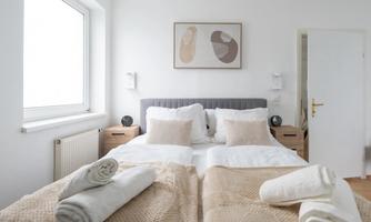 A modern bedroom with a large bed and beige bedding. The walls are light and there are large windows that let in natural light.