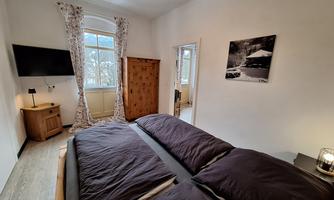 A cozy bedroom with a large bed and a television. Bright windows let in plenty of light and there is a nice wooden dresser.