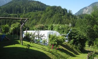 A green, hilly landscape with trees and mountains in the background. In the foreground, there is a greenhouse and colorful flowers.