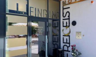 The entrance of a modern hotel with large windows and clear signage. In front of the door is a small wooden bench and a flower pot.