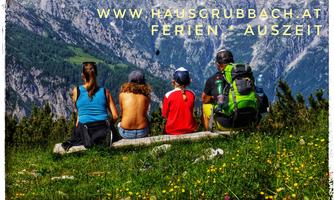 A group of people is sitting on a bench and looking at the mountains. The landscape is green with colorful flowers and tall mountains in the background.