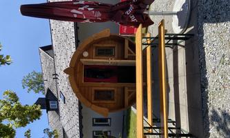A cozy wooden cabin with a round roof stands in front of a building. Wooden tables and chairs invite you to linger.