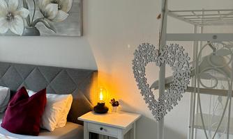 A cozy bedroom with a gray bed and colorful cushions. A heart-shaped coat rack hangs on the wall, and a cozy lamp provides warm light.