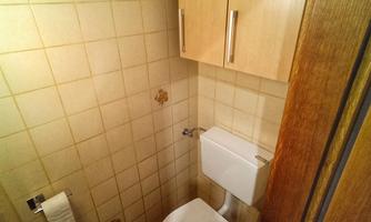 A small bathroom with a toilet, a sink, and a wooden cabinet. The walls are tiled, and the floor has a simple pattern.