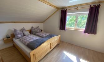 A cozy bedroom with a double bed and checkered bedspreads. The window lets in plenty of daylight, and the curtains are dark red.