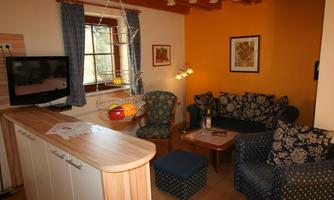 A cozy living room with a blue couch, an armchair, and a wooden table. In the background, there is a television, and the room is brightly lit.