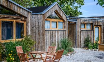 Modern wooden houses with large windows and an inviting courtyard. The area is surrounded by plants and offers a cozy spot to relax.