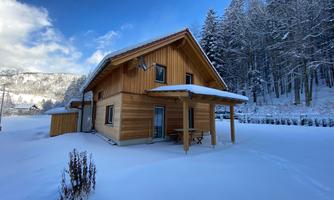 A cozy wooden house in the snow, surrounded by snow-covered trees. The sky is clear and shines in blue.