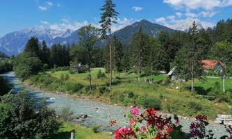 A scenic river flows through a green landscape with mountains in the background. Colorful flowers bloom in the foreground and the sky is blue with some clouds.