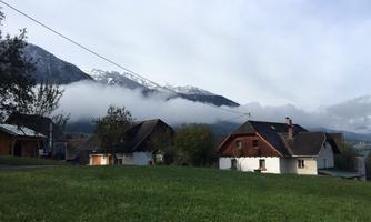 A tranquil rural scene with houses in a green meadow. In the background, there are mountains with clouds and snow.