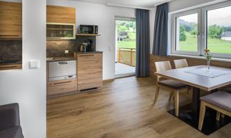 A modern kitchen with wooden details and a dining area. Large windows provide a view of the green landscape.
