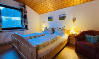 A cozy bedroom with a large double bed and a light wooden ceiling. Next to the bed is a comfortable armchair and there is a beautiful view through the window.