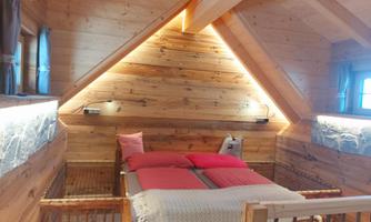 A cozy bedroom with a wooden frame bed and red pillows. The walls are wooden, and the light creates a warm atmosphere.