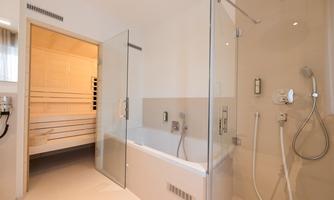 A modern bathroom with a glass shower and a bathtub. In the background, there is a wooden sauna.