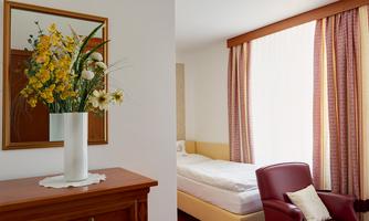 A cozy room with a single bed and a red armchair. A vase with yellow flowers sits on a dresser next to a mirror.