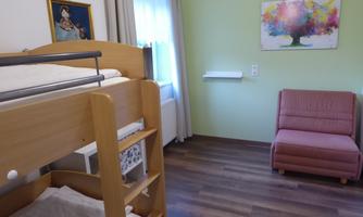 A bright room with a bunk bed and a cozy armchair. Colorful pictures hang on the walls and the floor is made of wood.