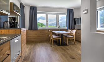 A modern kitchen with wooden surfaces and large windows. There is a dining table with chairs and a view of the mountains outside.