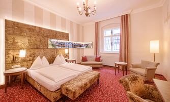A stylish hotel room with a double bed, elegant lighting, and comfortable seating. The walls are tastefully decorated, and there are large windows that let in natural light.