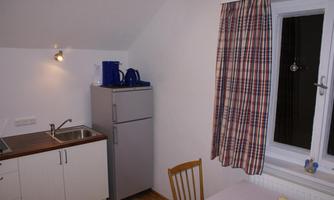 A small, bright kitchen unit with a refrigerator and a sink. Next to the window, there is a table with a chair and a tablecloth.
