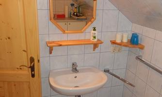 A small bathroom with a vanity, wooden shelves, and a mirror. The walls are tiled and there is a wooden stool.