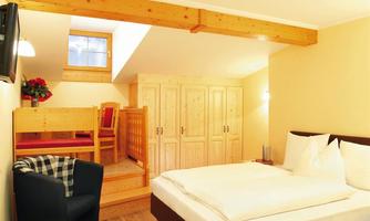 A cozy bedroom with a double bed and wooden paneling. In the background, there is a small dining area and a window that lets in natural light.