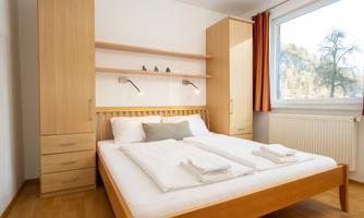 A modern bedroom with a double bed and light wooden furniture. The room has large windows that let in plenty of daylight.