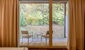 A cozy view of a terrace with two chairs and a table. The background shows an autumn forest with colorful leaves.