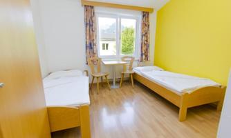 A simple room with two single beds and a table. The walls are painted yellow and there is a window with a view.