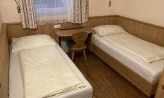 A simple room with two single beds and a small table. The walls are light, and the windows are furnished with curtains.