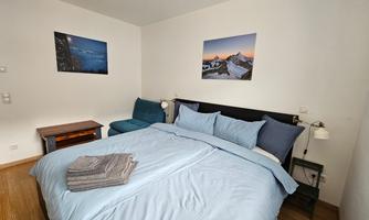 A cozy bedroom with a large bed and blue pillows. Pictures of landscapes hang on the walls, and the furniture is simple and modern.