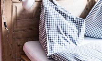 A cozy bedroom with a wooden frame and checkered pillows. A modern reading lamp hangs on the wall.