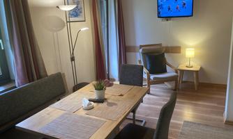 A cozy living area with a dining table, chairs, and a modern lamp. On the wall, a television is displaying a sports broadcast.