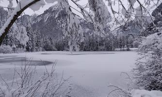 A snowy winter landscape with a frozen lake and snow-covered trees. The scene is quiet and peaceful, surrounded by mountains.
