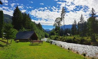 A picturesque landscape with a small wooden house by the river. Surrounded by trees and a clear sky.