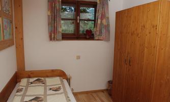 A simple bedroom with a single bed and a wooden wardrobe. The windows are adorned with colorful curtains and let light in.