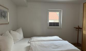 A simple bedroom with a double bed and white bedspreads. A window lets in daylight and there is a wooden shelf.