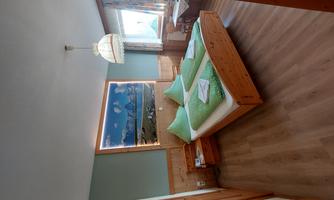 A cozy bedroom with a double bed and green pillows. There is a table with chairs and a large window that lets in plenty of light.