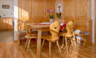 A cozy dining area with wood paneling and a light wood table. Flowers in a vase and a cross on the wall give the room an inviting atmosphere.