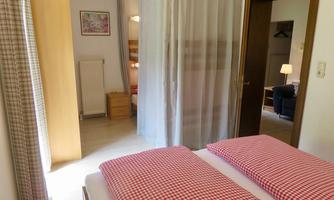 A cozy room with two beds that have red and white checked pillows. In the background, a closed door and a bright room with an armchair can be seen.