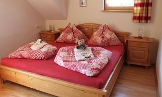 A cozy bedroom with a large wooden bed and red pillows. Next to the bed are two wooden nightstands and a window with checked curtains.