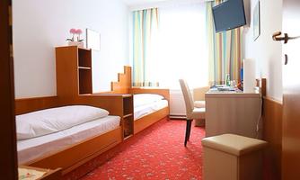 A simple room with two single beds and a small desk. A television hangs on the wall and light falls through a window with curtains.