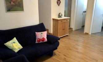 A cozy living room with a blue sofa and colorful cushions. The room has a wooden floor and bright walls.
