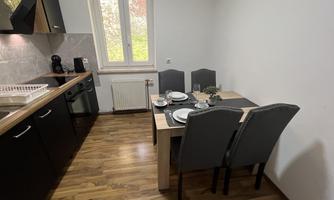 A modern kitchen with a dining table and chairs. The table is set and there are large windows that let in daylight.